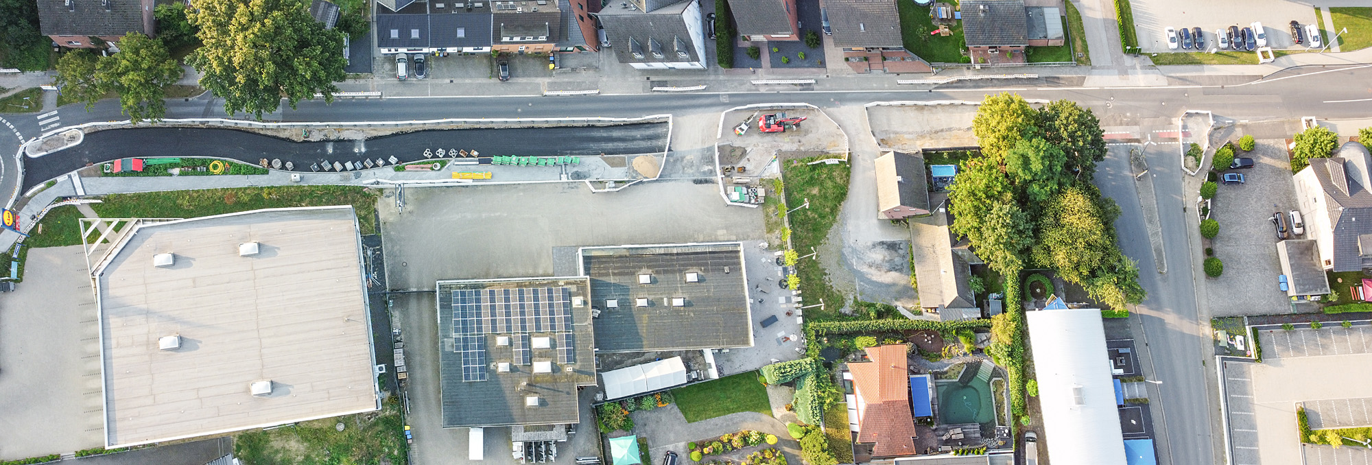 Luftaufnahme von oben (Vogelperspektive): unten Gebäude mit Sicht auf Dächer, mittig vor 2 Gebäuden Parkplatz, zwischen Gebäuden grüner Baum- & Pflanzenbewuchs & asphaltierte Straße, mittig 3 Baustellenbereiche mit Geräten auf Straße eingerahmt durch Schrankenzaun, linker Bereich mit neu asphaltierter Straße & Gehweg, 2 rechte Bereiche mit Boden bestehend aus Sand & Erde oben Häuser mit Sicht auf Dächer, dazwischen Grünflächen & grüner Baumbewuchs, oben & unten rechts Ausschnitt von Parkplätzen